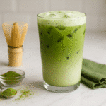 Starbucks matcha latte recipe - vibrant green iced matcha latte in clear glass with bamboo whisk and matcha powder on white marble counter